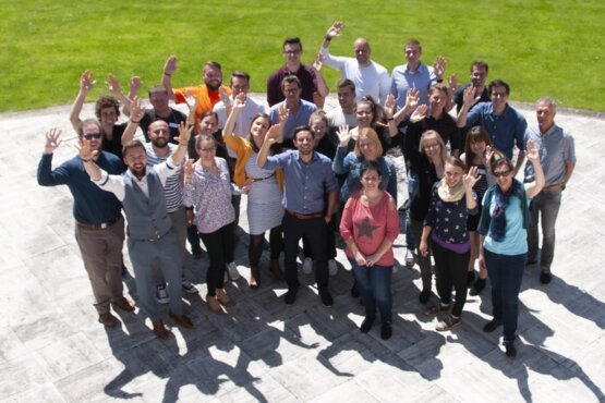 Gruppe von Menschen vor einer grünen Wiese von oben mit Blick in die Kamera
